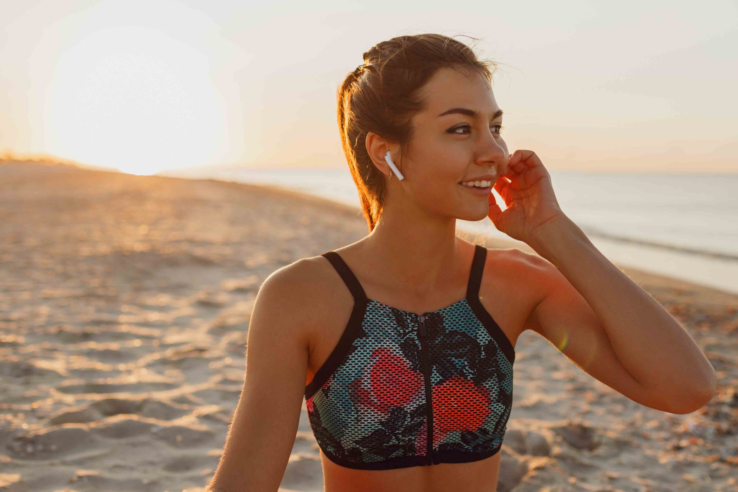 Smiling young female  listening music in headphones after training. Young athletic woman exercising near the sea . Summer sunset.