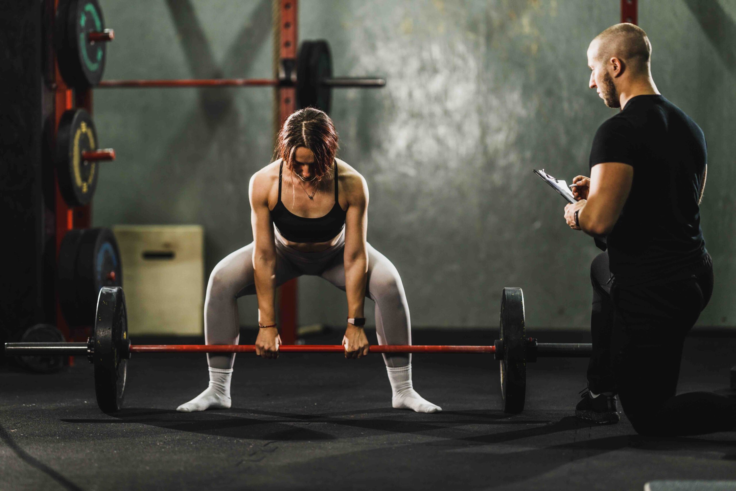 Personal trainer watching and assisting a muscular young woman in weight training at the gym. She is doing deadlift exercise with barbell.