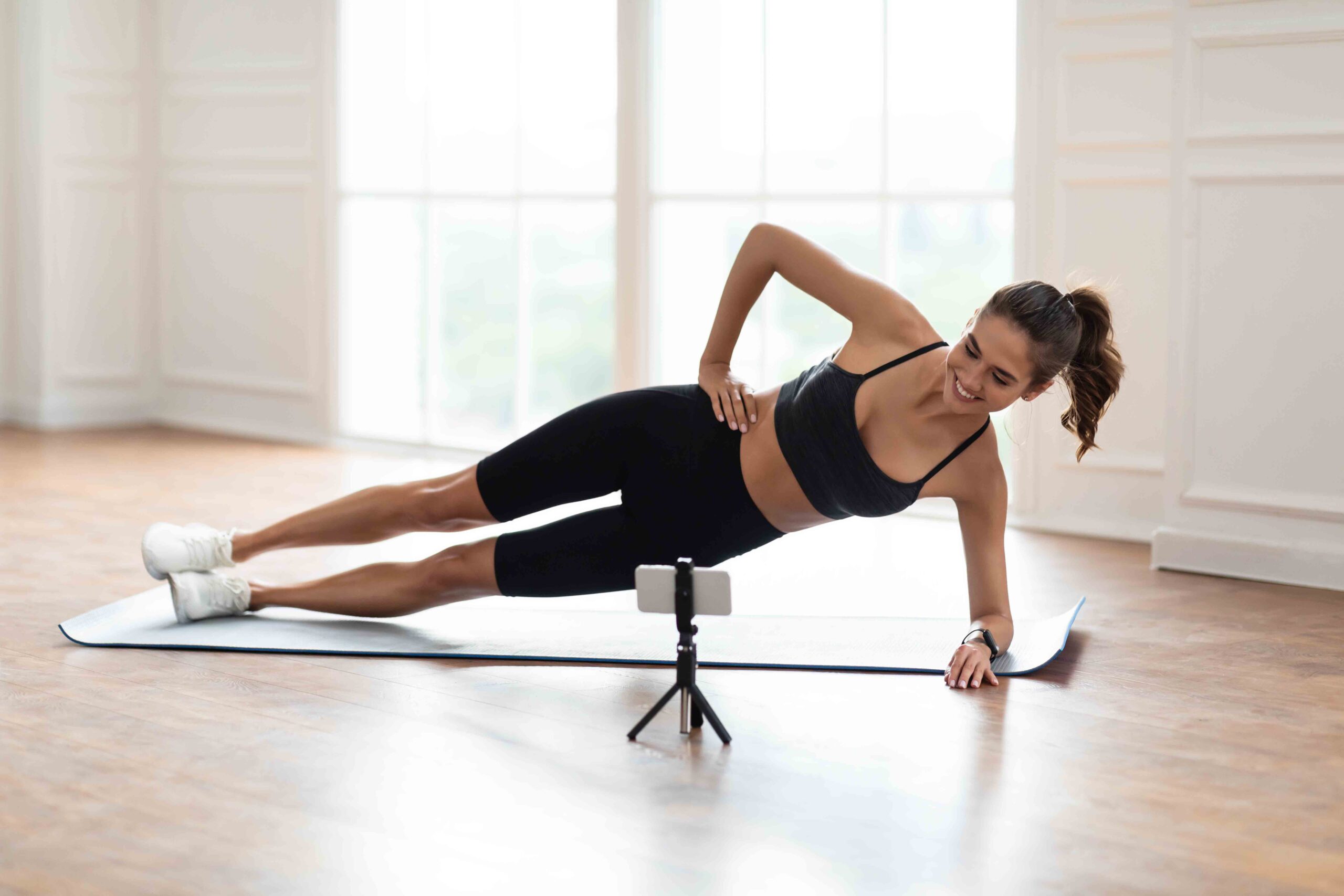Online Workout. Smiling Sporty Female Standing In Side Plank Position On Yoga Mat Near Window, Looking At Cellphone On Tripod, Watching Tutorial Or Recording Video For Blog, Having Chat With Coach